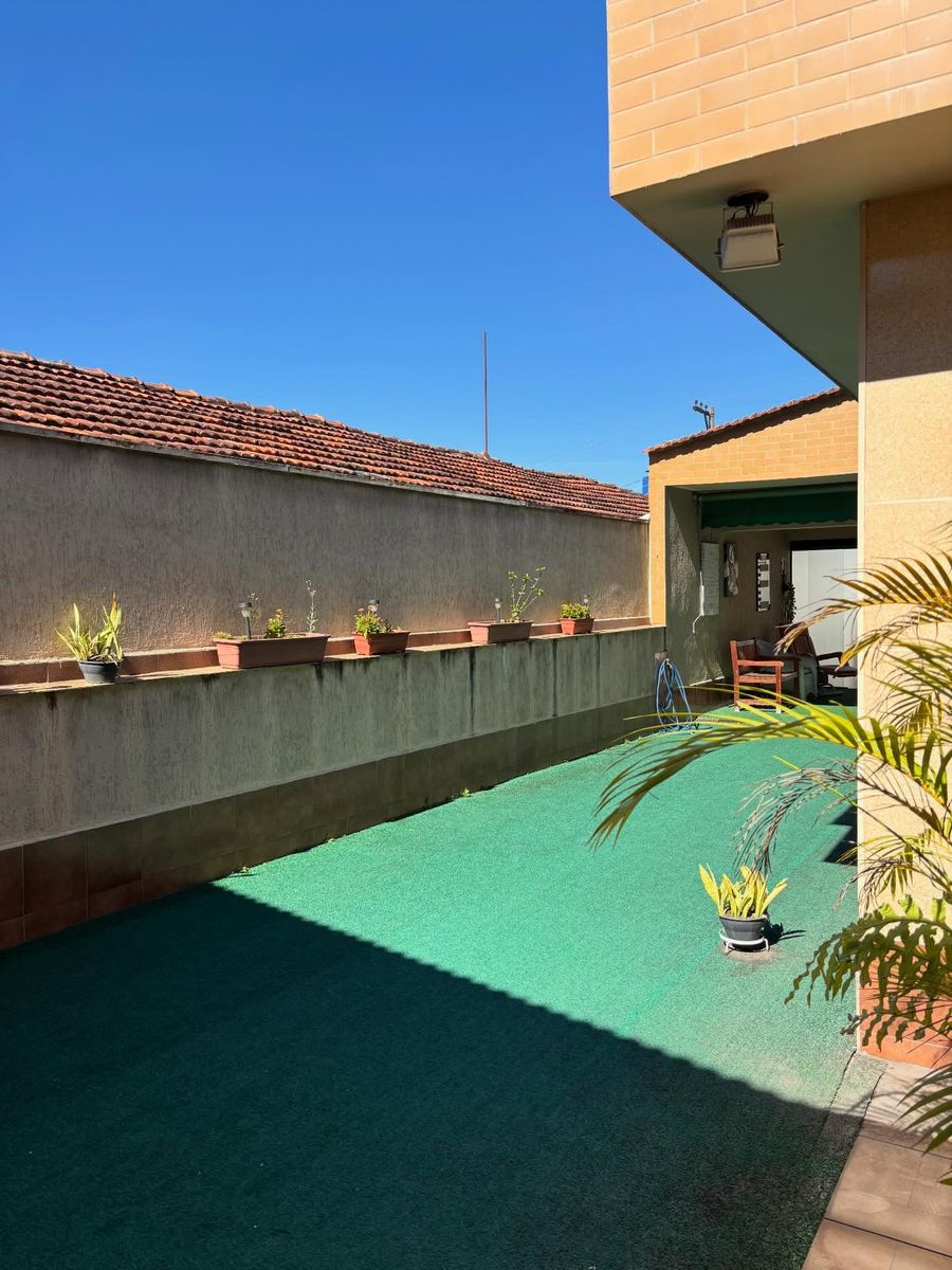 Área externa ensolarada com piso de grama sintética, muro baixo com uma fileira de floreiras e luminárias, e ao fundo a área coberta com banco de madeira e vasos de palmeira.