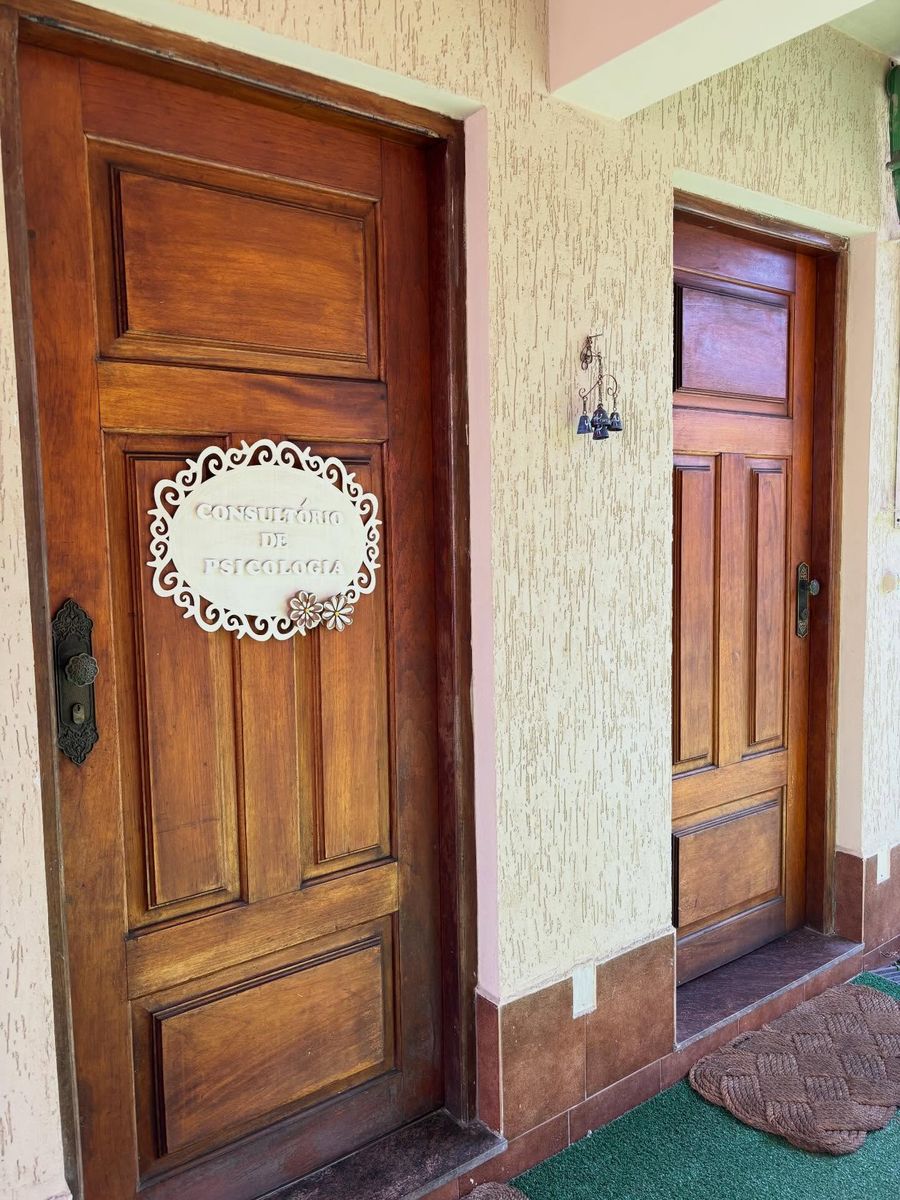 Duas portas de madeira maciça lado a lado, sob a área coberta da entrada. Na porta da esquerda, uma placa oval branca vazada com a inscrição Consultório de Psicologia.