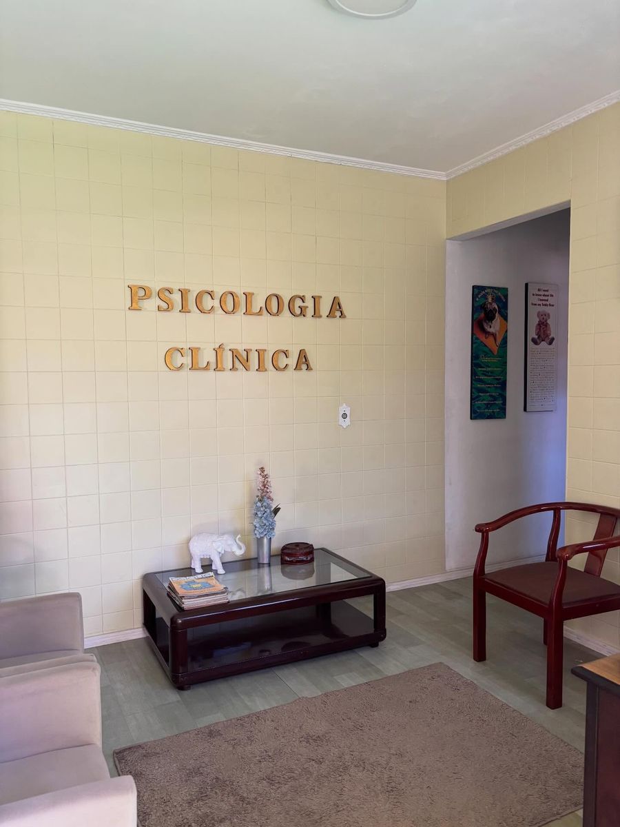 Sala de espera com sofá claro, mesa de centro baixa com revistas e um vaso de flores secas, poltrona de madeira e tapete. Na parede, em letras douradas, Psicologia Clínica.