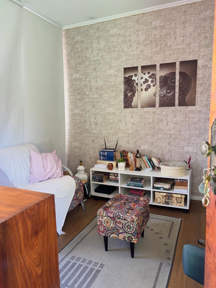 Sala de atendimento com parede de tijolo branco, sofá claro com almofada, estante baixa cheia de livros, banqueta estampada sobre um tapete e um quadro em quatro partes com dois perfis humanos formados por engrenagens.