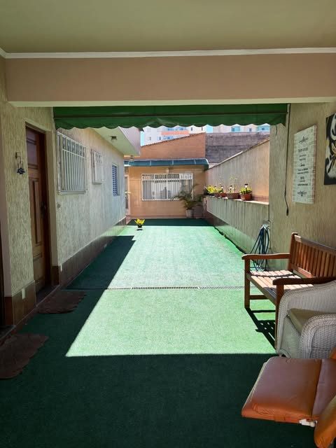 Varanda coberta com piso de grama sintética e toldo verde, com banco de madeira e poltronas encostados na parede, aberta para o corredor externo com vasos de plantas e a fachada da casa ao fundo.